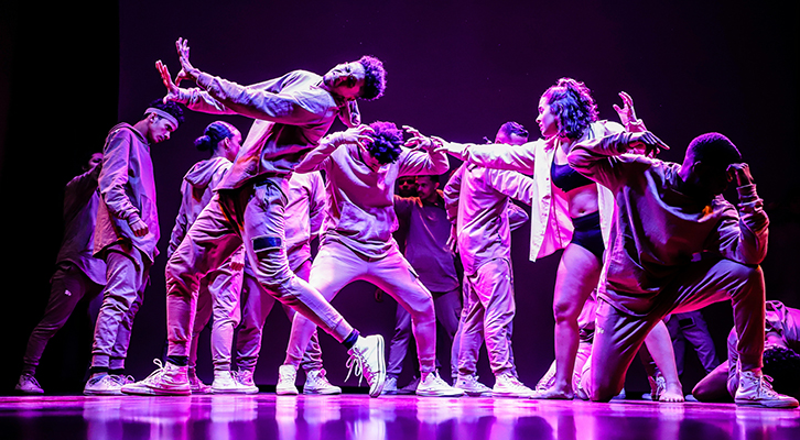 A group dancers striking dynamic poses on stage under vibrant purple lighting