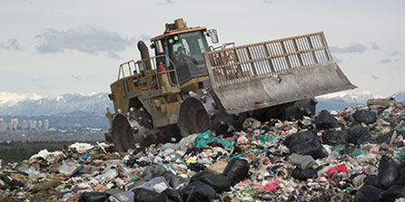 A bulldozer drives over a large mound of garbage at the Vancouver Landfill with the North Shore Mountains in the background on a cloudy day