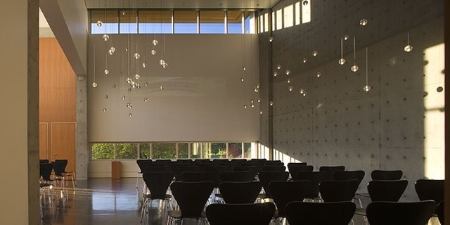 Interior of a modern hall with high ceilings, large windows allowing natural light, suspended decorative lighting, and rows of black chairs arranged for a gathering.