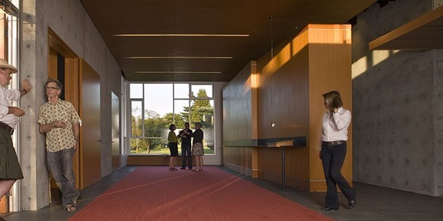 Reception area with warm wooden accents and a red carpet, where several people are casually gathered, talking near large windows that let in natural light