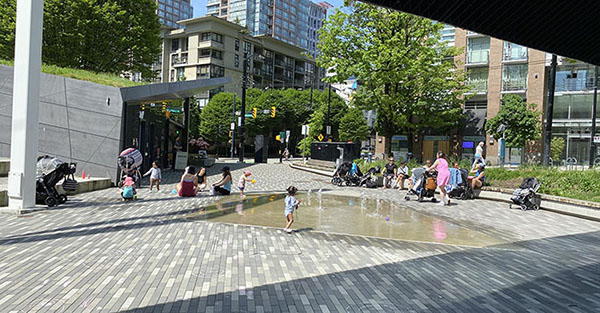 Children playing in a splash pad while parents and caregivers watch from the edge on a sunny day.