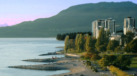 View of Sunset Beach in Vancouver with waterfront buildings, trees, and mountains in the background at dusk.