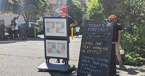 People stand beside a chalkboard sign showing a heat warning and cooling centre hours in a West End neighbourhood.