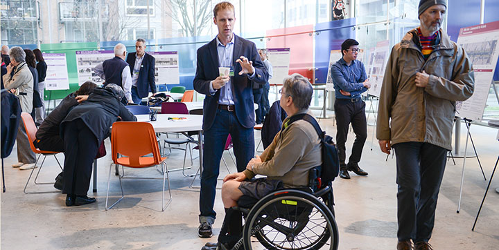 A person in a wheelchair speaking with a planner at an open house with people talking at tables and viewing display boards in the background A person in a wheelchair speaking with a planner at an open house with people talking at tables and viewing display boards in the background