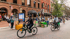 Peple hanging around Water St in Gastown. 2 cyclists ride on the road. In the back a group of people are sitting around a bench.