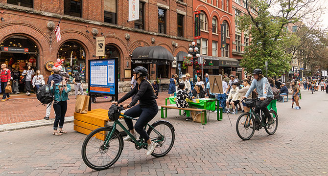 Peple hanging around Water St in Gastown. 2 cyclists ride on the road. In the back a group of people are sitting around a bench.