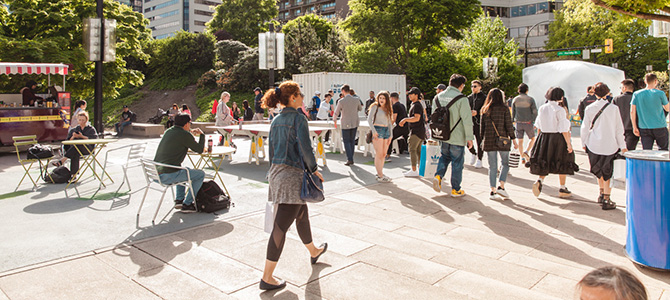People gathering at tables and walking through the 800 Robson public plaza.