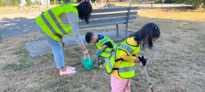 One adult and two children in high-visibility vests using litter pickers and a bucket to clean up grass in a park. 