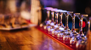 Empty wine glasses lined up on a bar counter, with a blurred background featuring dimly lit bar ambiance.