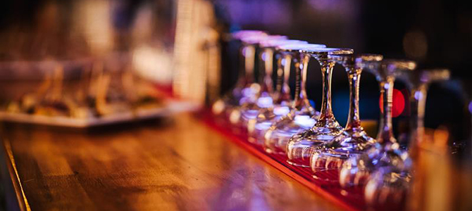 Empty wine glasses lined up on a bar counter, with a blurred background featuring dimly lit bar ambiance.