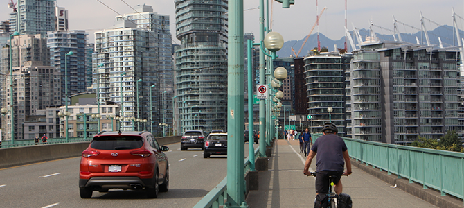 	A cyclist on the Cambie Bridge bike lane alongside cars on a bridge with city buildings in the background.