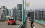 A cyclist on the Cambie Bridge bike lane alongside cars on a bridge with city buildings in the background.