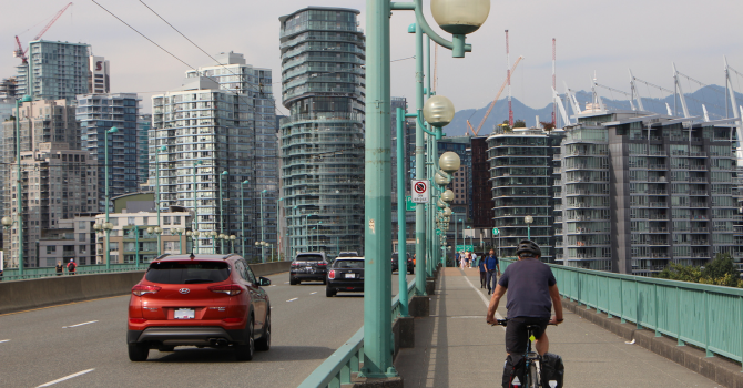 A cyclist on the Cambie Bridge bike lane alongside cars on a bridge with city buildings in the background.