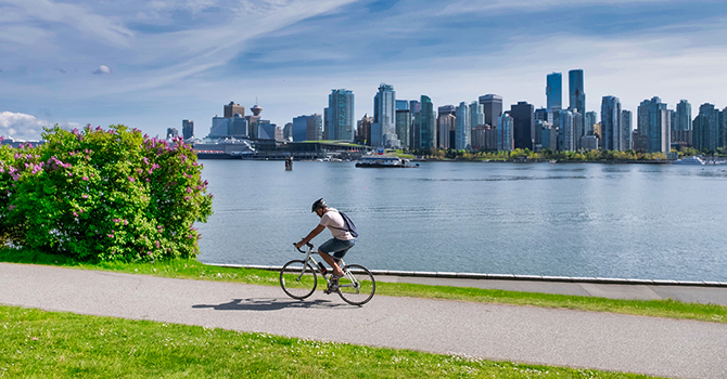 Person cycling along a waterfront bike path with the Vancouver skyline in the background on a sunny day