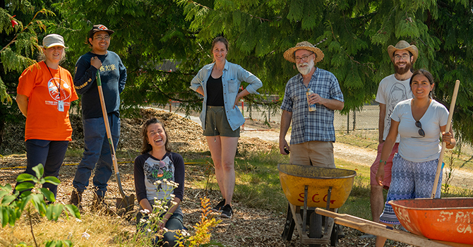 Group of gardeners, some holding shovels, standing next to wheelbarrows among cedar trees in a sunlit garden.