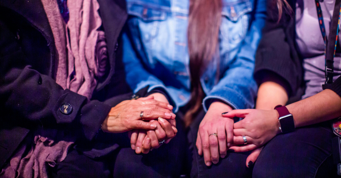 Hands of three people sitting side by side, holding each other’s hands in support.
