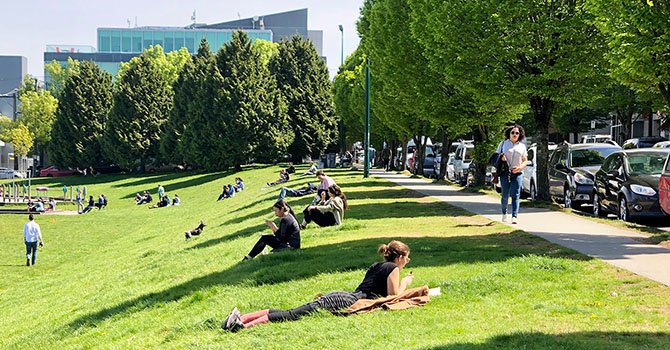 People relaxing on a grassy hill in a park on a sunny day, with trees lining a sidewalk and parked cars along the street. People relaxing on a grassy hill in a park on a sunny day, with trees lining a sidewalk and parked cars along the street.