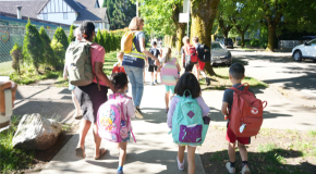Kids and adults walking on a sidewalk through a sunny neighbourhood
