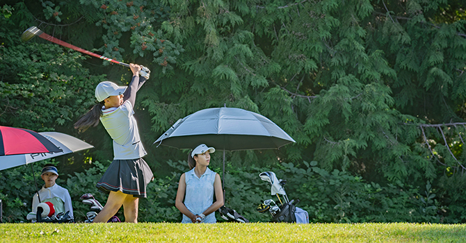 Female golfer executing a powerful swing on a sunny course, observed by onlookers and a caddy, with lush greenery in the background.