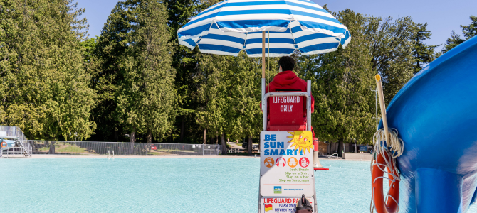 Lifeguard sitting in a chair beside an outdoor pool with a blue umbrella overhead
