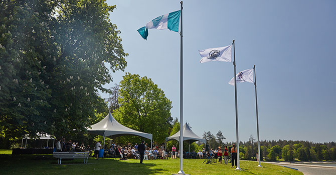MST flag raised on flagpoles during a community gathering with attendees seated under tents in a park.
