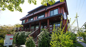 Large red and black multiplex home viewed from one corner of an intersection, mailbox visible outside the front steps and vegetation on all sides of the property