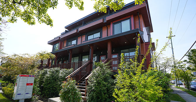 Large red and black multiplex home viewed from one corner of an intersection, mailbox visible outside the front steps and vegetation on all sides of the property