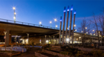 NEU building under the Cambie Bridge at dusk, with illuminated exhaust stacks and raised garden beds in the foreground.