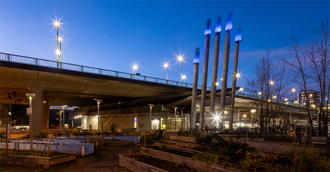 NEU building under the Cambie Bridge at dusk, with illuminated exhaust stacks and raised garden beds in the foreground. NEU building under the Cambie Bridge at dusk, with illuminated exhaust stacks and raised garden beds in the foreground.