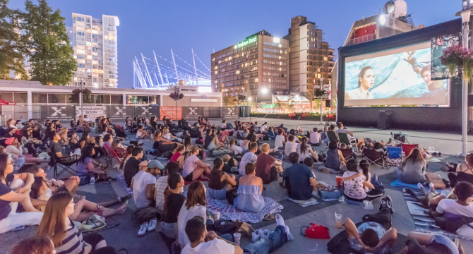 Crowd of people sitting on blankets and chairs at an outdoor movie screening with BC place and buildings in the background.