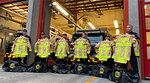 A group of firefighters standing in front of a firetruck, each holding new yellow turnout jackets labeled 'Vancouver Fire.' Black gear bags are placed on the ground in front of them.