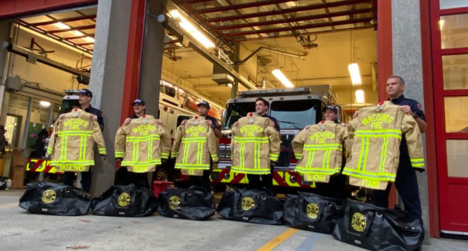 A group of firefighters standing in front of a firetruck, each holding new yellow turnout jackets labeled 'Vancouver Fire.' Black gear bags are placed on the ground in front of them.