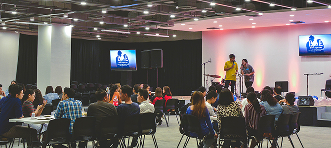 An indoor pop-up event with two speakers on the stage and groups of attendees sitting at tables.