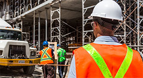 3 workers at a constuction site, wearing hard hats and reflective vests. A large cement track is parked close to them.