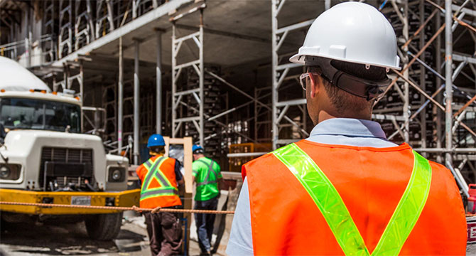 3 workers at a constuction site, wearing hard hats and reflective vests. A large cement track is parked close to them.