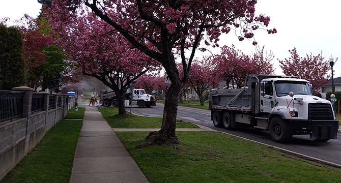 Two City cement trucks on a residential street. One truck is pouring concrete and workers are spreading it on the road. 