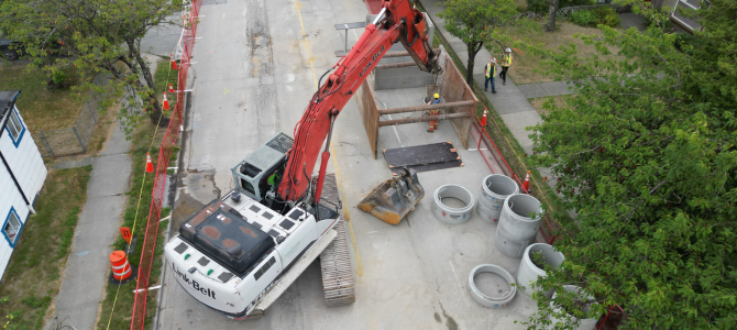 Aerial view of excavator on a city street with workers and concrete pipe sections on site.