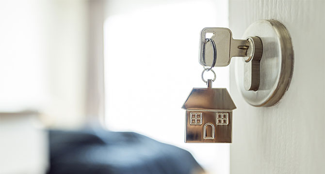 An open door leading to a room. On the door lock is a key with a keychain in the shape of a house. 