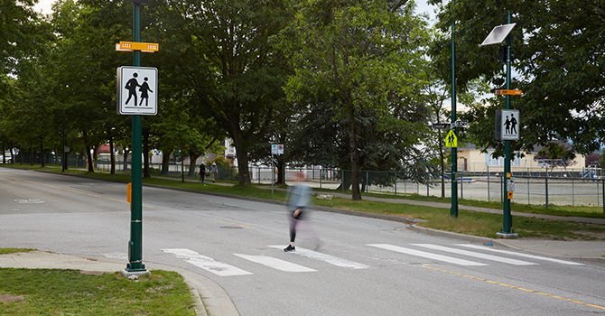 Pedestrian crossing a marked crosswalk on a tree-lined street, with flashing amber lights and school crossing signs visible.
