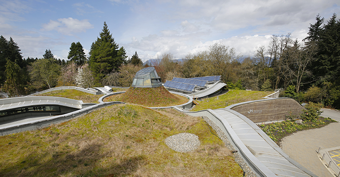 View of VanDusen Botanical Garden's visitor center with green roofs, featuring grassy areas and integrated solar panels, surrounded by diverse trees with a backdrop of distant mountains.