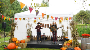 Two musicians playing violin and guitar on an outdoor stage decorated with pumpkins, cornstalks, flowers, and colorful bunting under a white canopy.