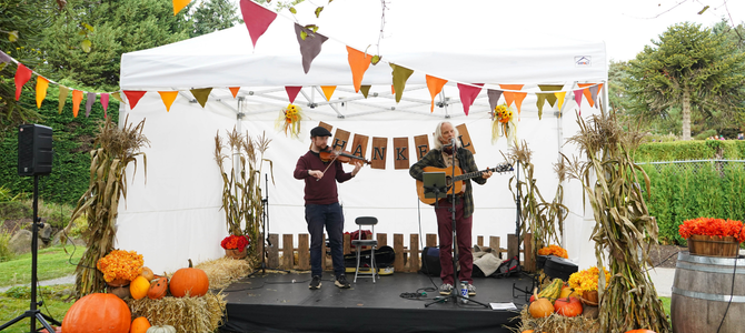 Two musicians playing violin and guitar on an outdoor stage decorated with pumpkins, cornstalks, flowers, and colorful bunting under a white canopy.