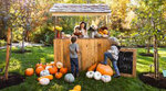 A woman stands at a wooden booth holding a toddler, while two other young children talk to her, standing among pumpkins of different sizes.