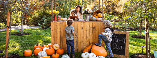 A woman stands at a wooden booth holding a toddler, while two other young children talk to her, standing among pumpkins of different sizes.