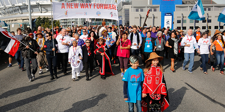 A crowd of people walking in the 2017 Walk for Reconciliation on the Georgia Viaduct, some holding a banner reading 'A new way forward', some waving flags, some wearing Reconciliation Canada t-shirts, with First Nations elders and children in regalia