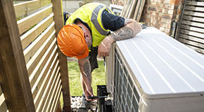 Worker in a safety vest and hard hat installing or servicing an outdoor heat pump unit.