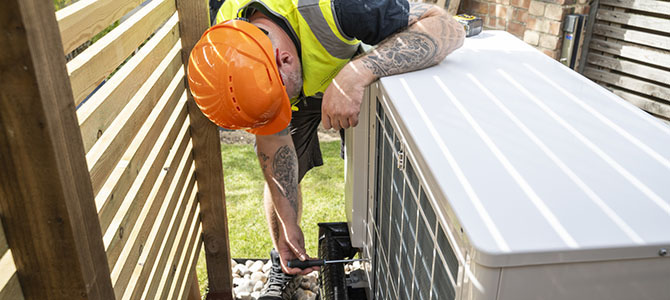 Worker in a safety vest and hard hat installing or servicing an outdoor heat pump unit. Worker in a safety vest and hard hat installing or servicing an outdoor heat pump unit.