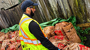 Worker wearing a high-visibility vest and gloves stands beside large paper yard waste bags near a wooden fence.