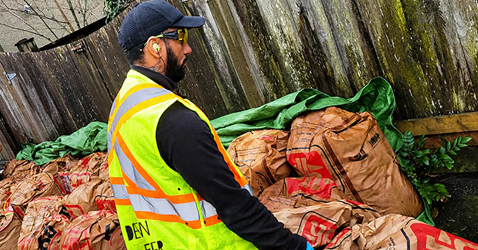 Worker wearing a high-visibility vest and gloves stands beside large paper yard waste bags near a wooden fence.