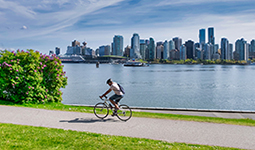 Person cycling along a waterfront bike path with the Vancouver skyline in the background on a sunny day